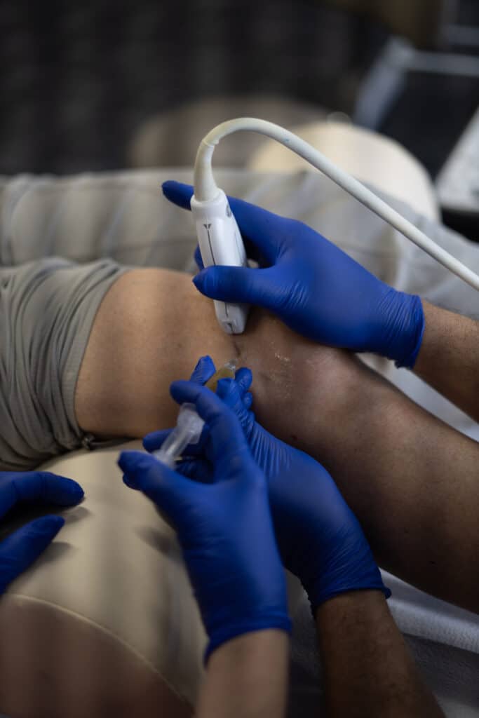 Ultrasound-guided knee injection: gloved hands hold the probe and the syringe at the knee