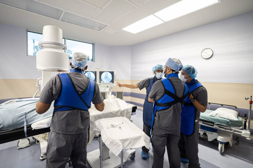 Clinicians in lead aprons review fluoroscopy images beside a procedure table