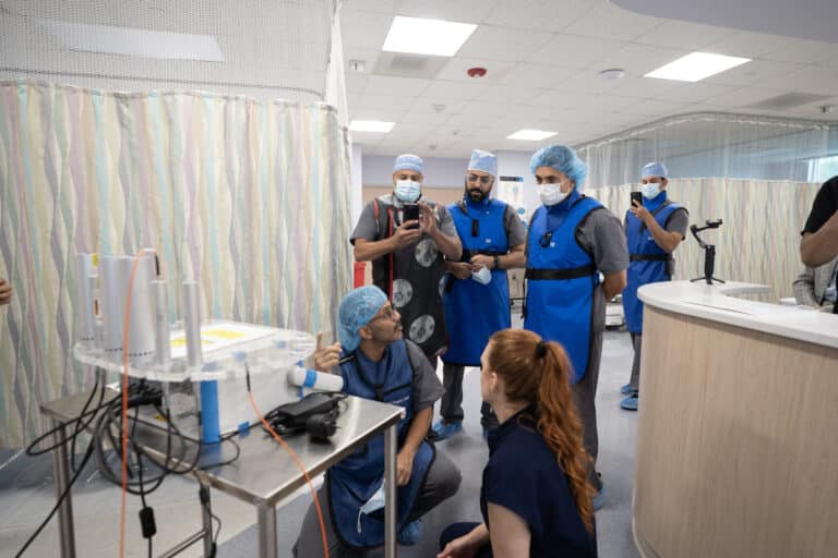 A clinician in a lead apron kneels at an equipment cart and explains the setup while trainees look on