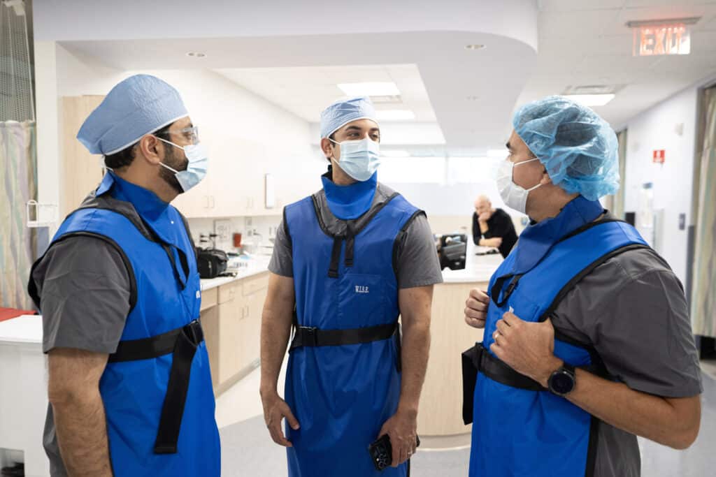 Our Story 5 Three clinicians in lead aprons and surgical caps talk in a procedure suite