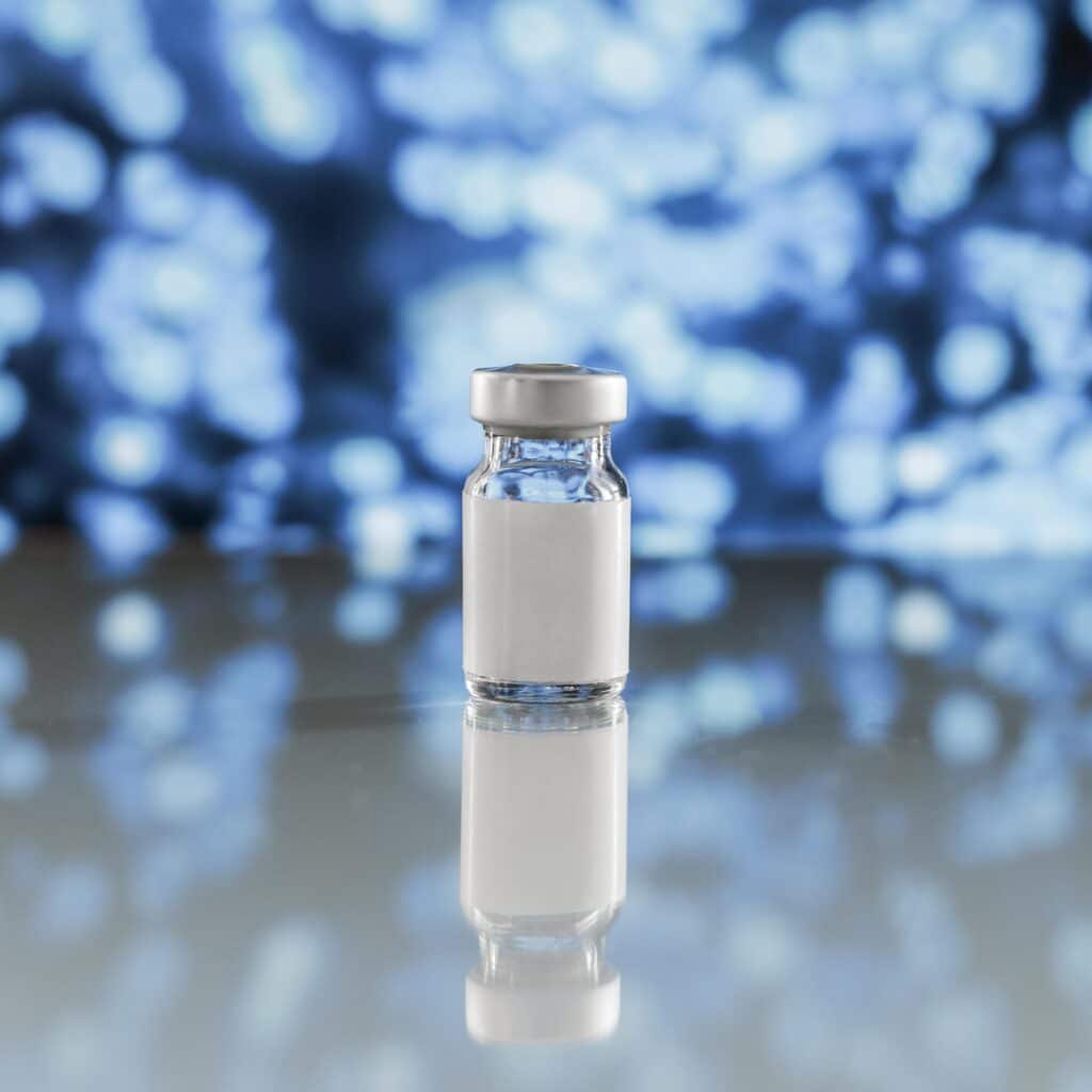 Home 2026 9 A small glass vial with a silver cap on a reflective surface against a blurred blue background